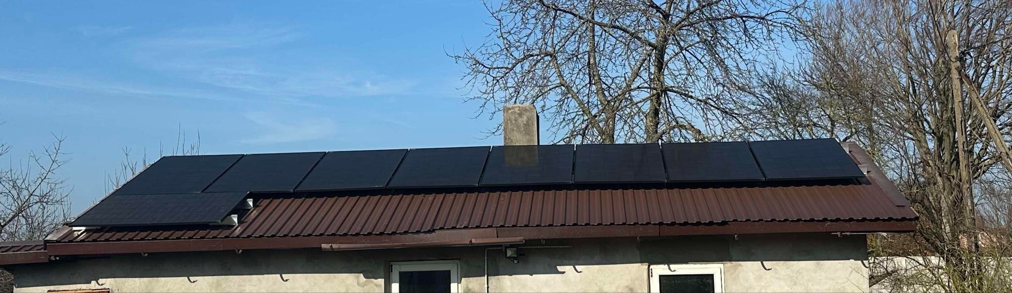 Panele fotowoltaiczne na brązowym dachu domu jednorodzinnego w słoneczny dzień. Widoczny komin i drzewa w tle. Instalacja paneli fotowoltaicznych.