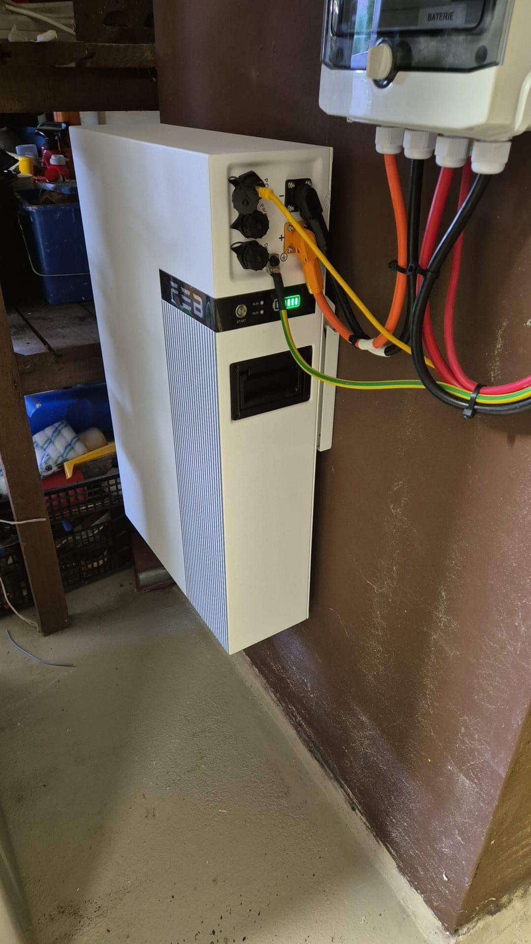 Biały magazyn energii z podłączonymi kablami, widoczne diody LED i złącza. Instalacja w pomieszczeniu o brązowych ścianach i szarej podłodze.