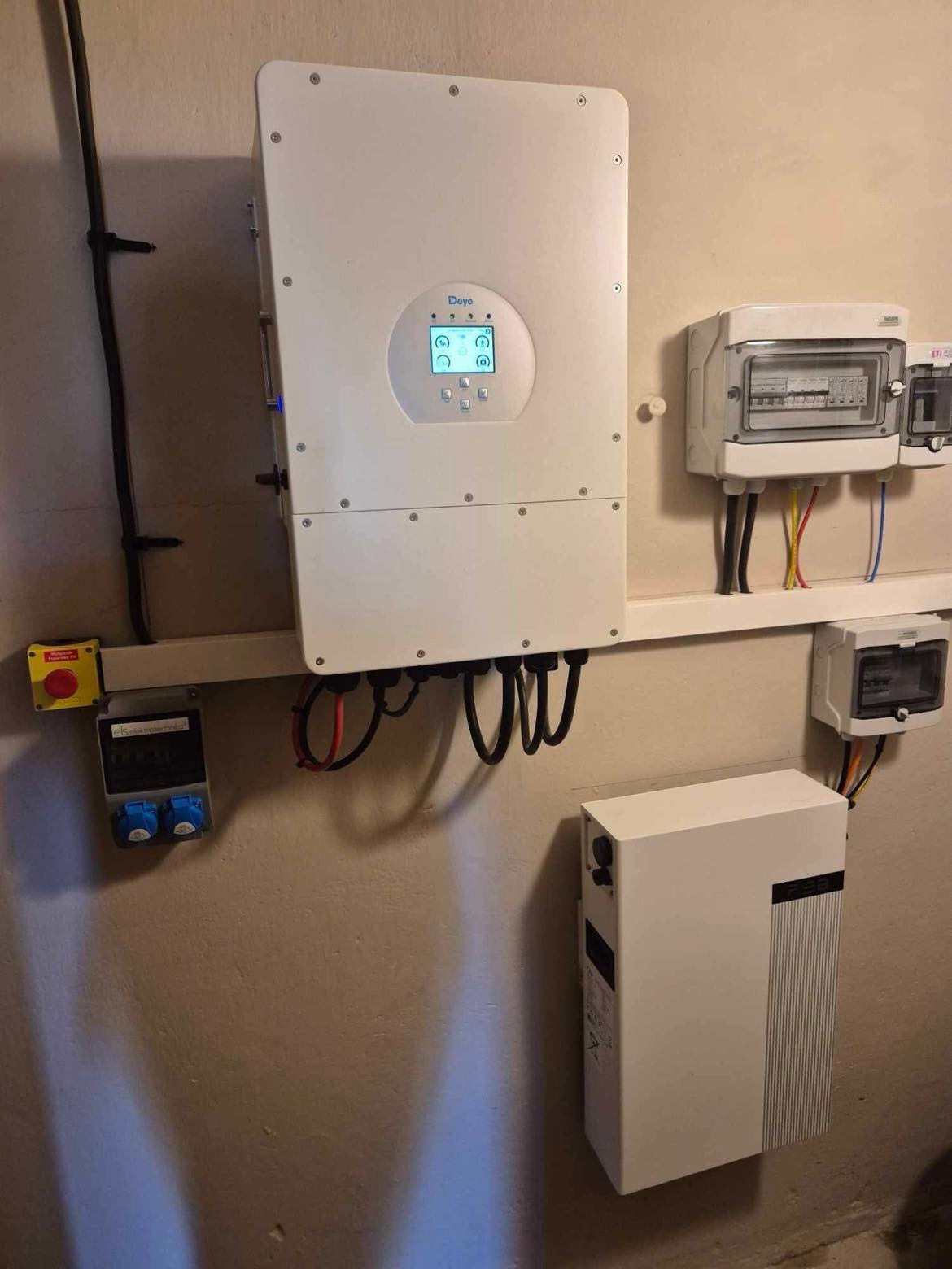 Biały inwerter fotowoltaiczny Deye zamontowany na ścianie, z widocznymi kablami, skrzynką bezpiecznikową i akumulatorem energii. Instalacja w pomieszczeniu technicznym.