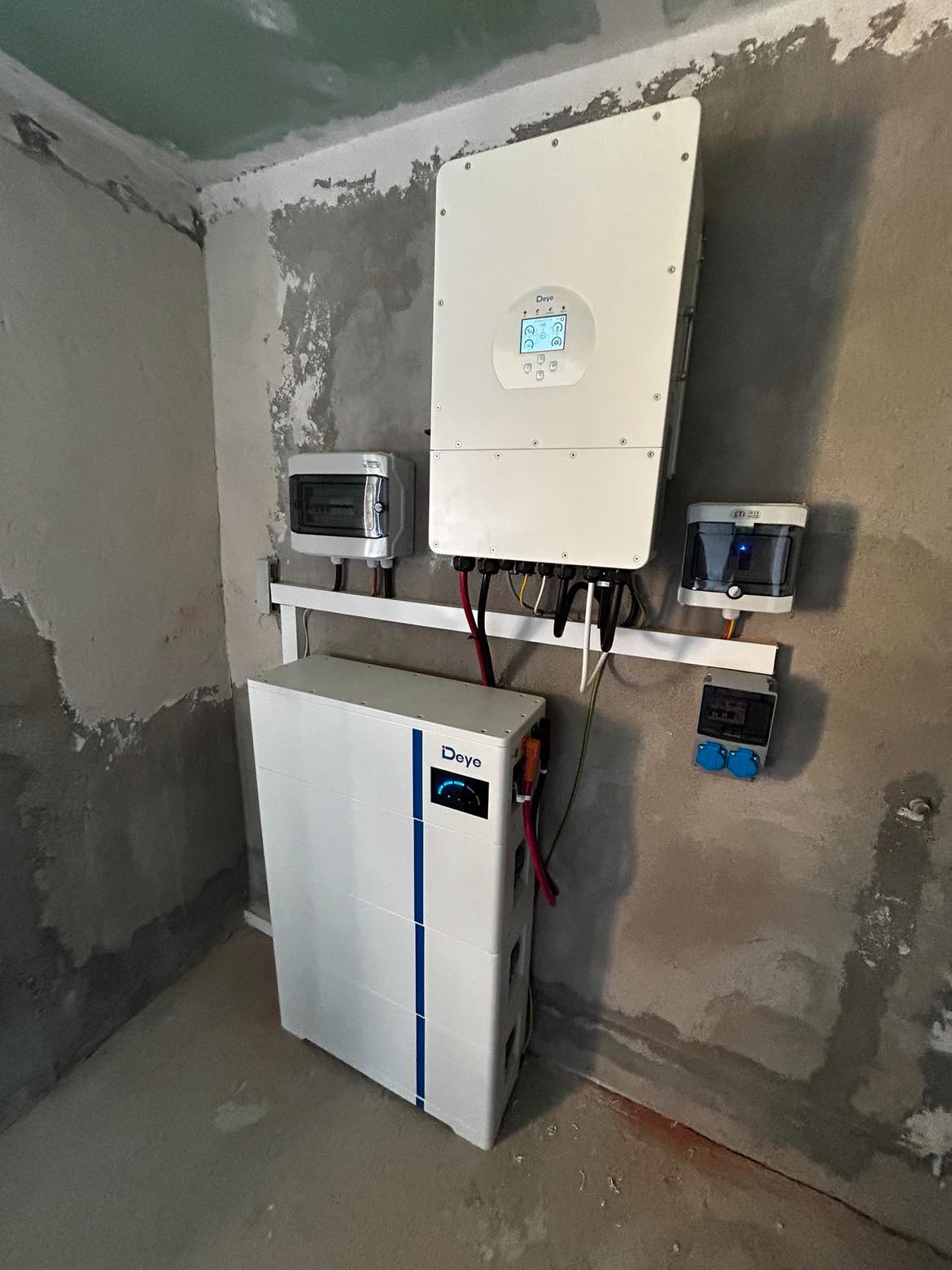 Biały magazyn energii Deye z inwerterem i rozdzielnicami na surowej ścianie. Widoczne kable i elementy instalacji. Montaż w trakcie wykończenia.