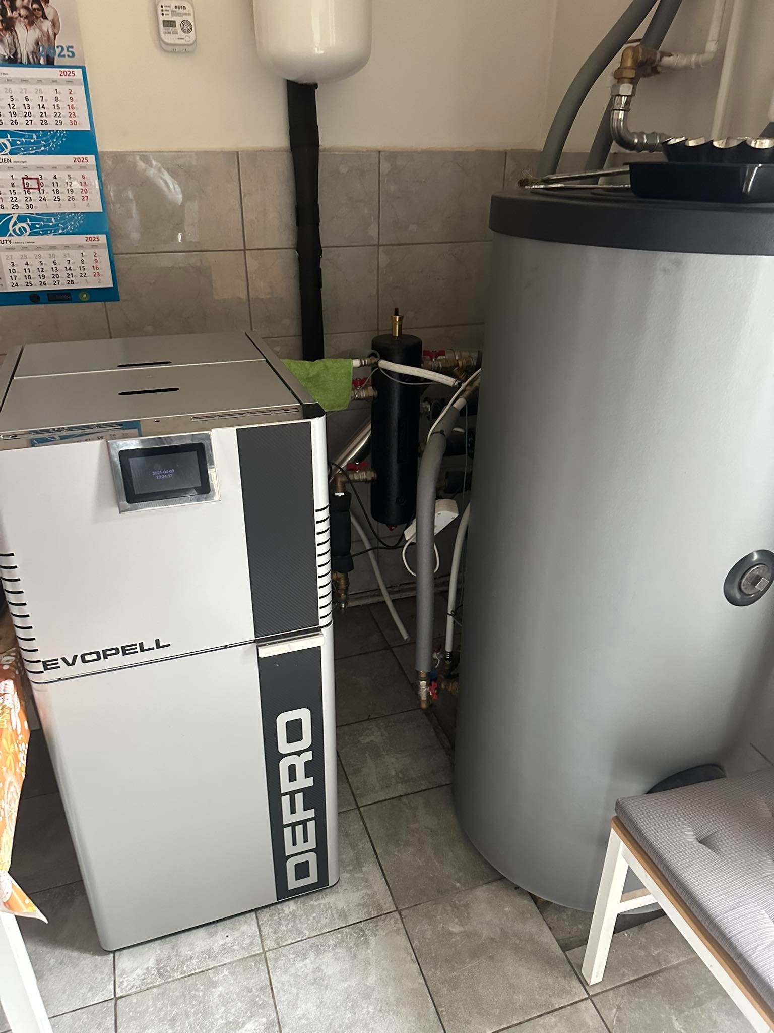 Kompaktowy piec na pellet marki Defro Evopell obok szarego zasobnika ciepłej wody w kotłowni z kalendarzem na ścianie. Widoczne rury i instalacja.