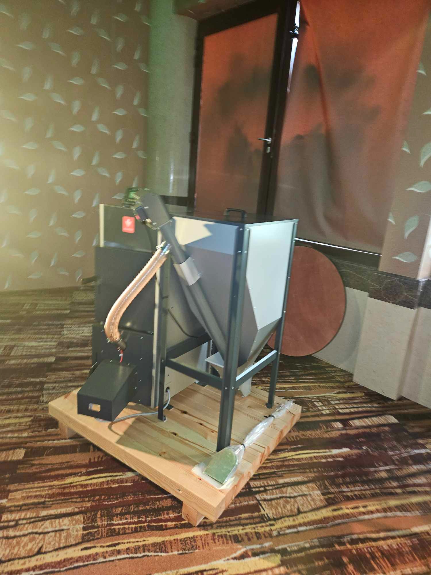 Nowoczesny piec na pellet z podajnikiem, ustawiony na drewnianej platformie w pomieszczeniu z wzorzystą tapetą. Widoczne rury i elementy konstrukcyjne.