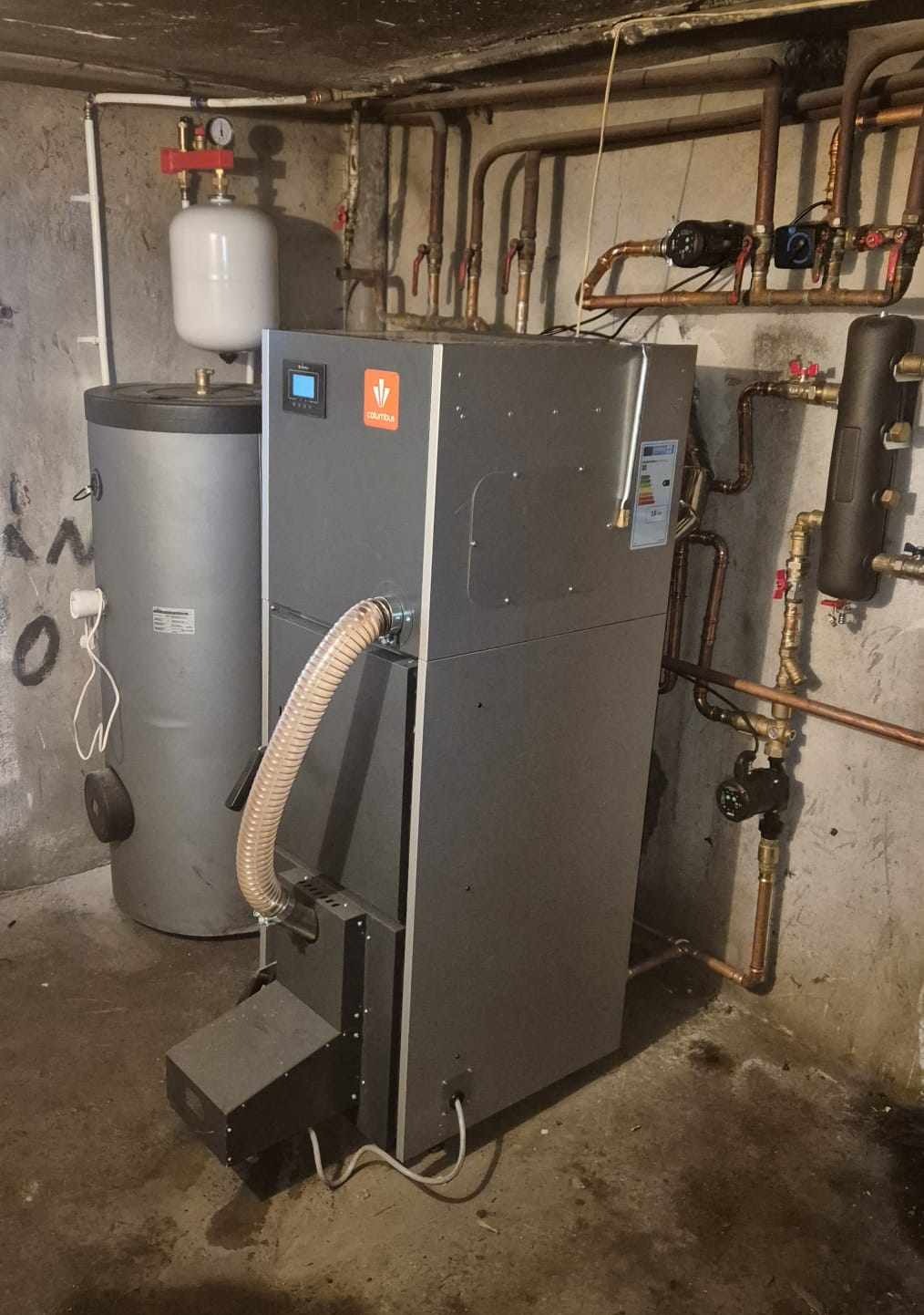 Szary piec Columbus z podajnikiem i zbiornikiem na wodę w kotłowni. Widoczne rury instalacji grzewczej i manometr. Oświetlenie górne.