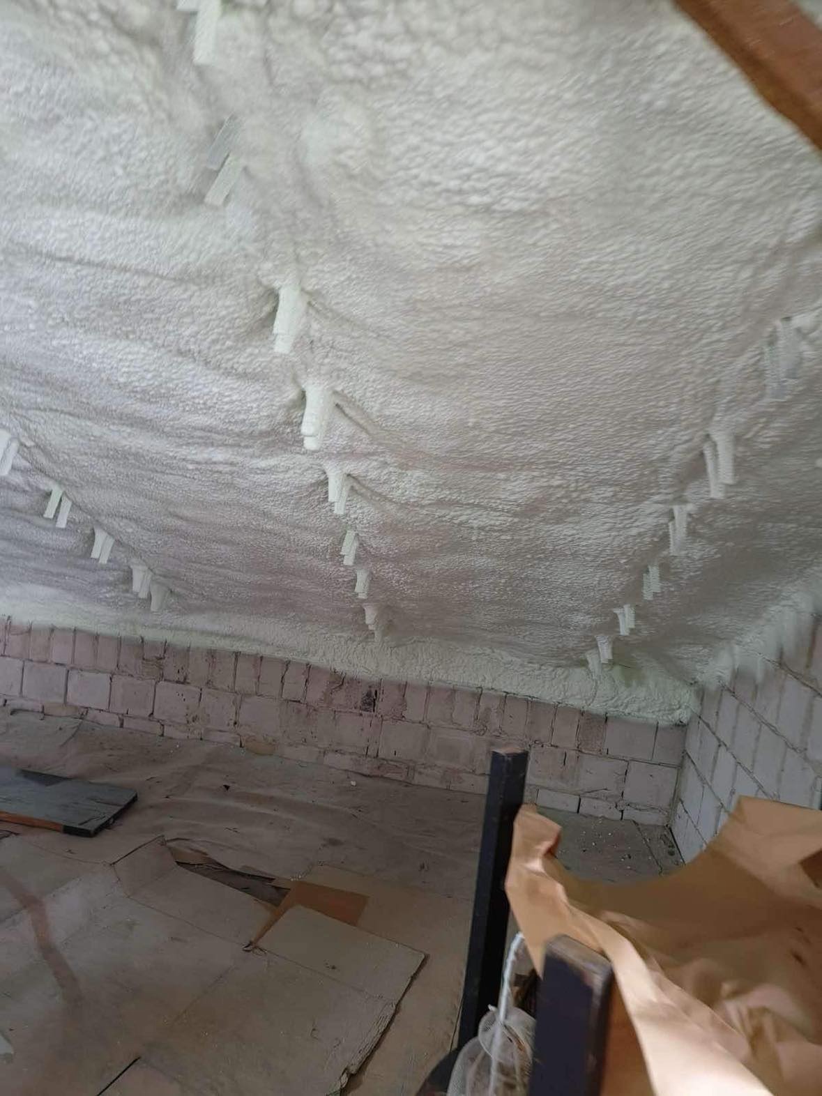 Izolacja poddasza pianką PUR z widocznymi plastikowymi kołkami mocującymi. Biała, nierówna powierzchnia piany pokrywa całą przestrzeń. Widoczne fragmenty muru i podłogi.