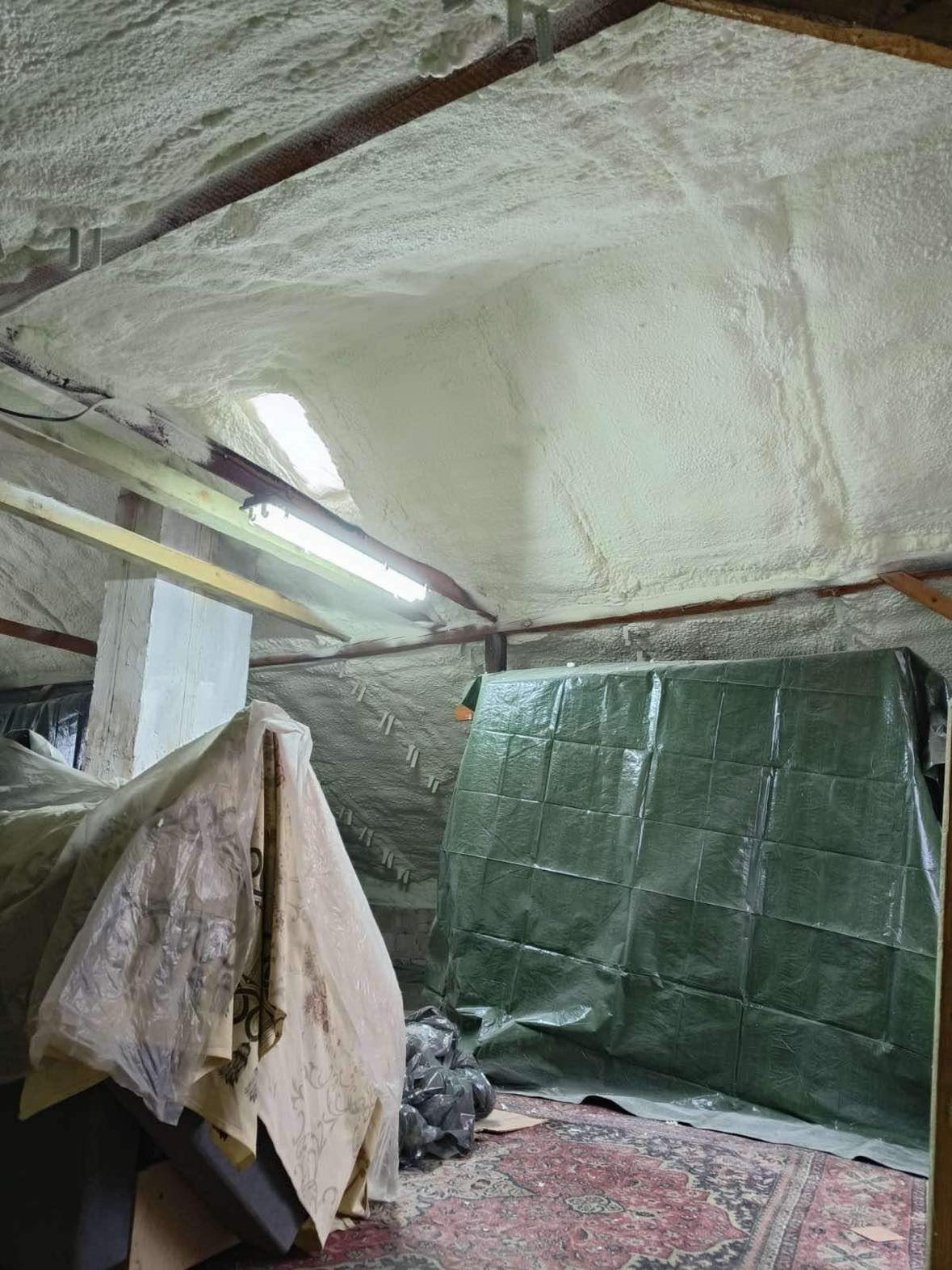Izolacja poddasza pianą PUR, widoczne pokryte pianą krokwie i skosy. Część pomieszczenia zabezpieczona folią. Oświetlenie jarzeniowe.