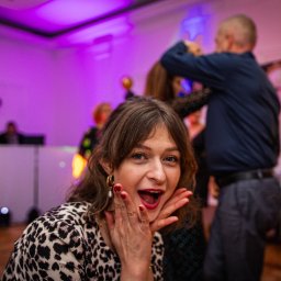 Patryk Fijołek - Rozradowana kobieta w bluzie w panterkę na imprezie, z otwartymi ustami i dłońmi przy twarzy. W tle DJ i tańczący ludzie, fioletowe oświetlenie.