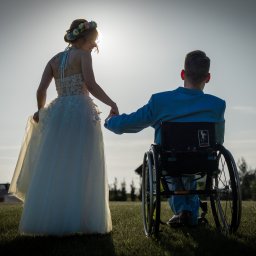 Patryk Fijołek - Para młoda na tle nieba: panna młoda w sukni ślubnej trzyma za rękę pana młodego na wózku inwalidzkim. Ujęcie z tyłu, oświetlenie kontrowe.