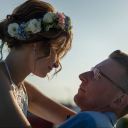 Patryk Fijołek - Zakochana para w zbliżeniu: panna młoda z wiankiem i pan młody w okularach patrzą na siebie w słoneczny dzień. Romantyczny moment.