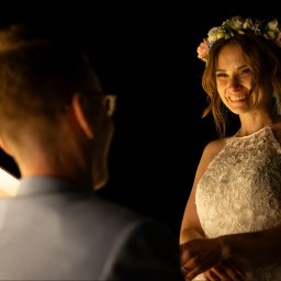 Patryk Fijołek - Uśmiechnięta panna młoda w wianku i koronkowej sukni, spogląda na mężczyznę w okularach, oświetlona ciepłym światłem na ciemnym tle.