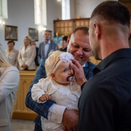 Patryk Fijołek - Uśmiechnięty mężczyzna w garniturze trzyma na rękach niemowlę w białej sukience i opasce z kwiatkiem, podczas uroczystości w kościele, w tle widoczni goście.