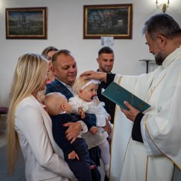 Patryk Fijołek - Ksiądz udziela sakramentu chrztu dziewczynce w kościele. W tle rodzina dziecka. Uroczystość w jasnym wnętrzu z obrazami na ścianach.