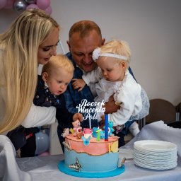 Patryk Fijołek - Rodzina świętuje pierwsze urodziny dziecka przy torcie z napisem 'Roczek Wincenta Poli', dekorowanego motylami i misiami. Dzieci obserwują płomień świeczki.