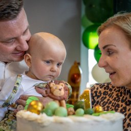 Patryk Fijołek - Rodzinne świętowanie: tata całuje rocznego synka trzymanego nad tortem z lwem i świeczką numer 1, mama patrzy z uśmiechem. Tło: zielone balony.