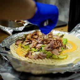 Patryk Fijołek - Przygotowywanie tortilli z mięsem, sałatą i sosem na folii aluminiowej. Dłoń w niebieskiej rękawiczce dodaje składniki. Kulinarna fotografia produktowa.