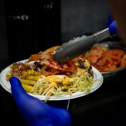 Patryk Fijołek - Nakładanie szczypcami pomidora na porcję frytek, surówki i mięsa na papierowym talerzu, trzymanym w dłoni w niebieskiej rękawiczce, w ciemnym otoczeniu.