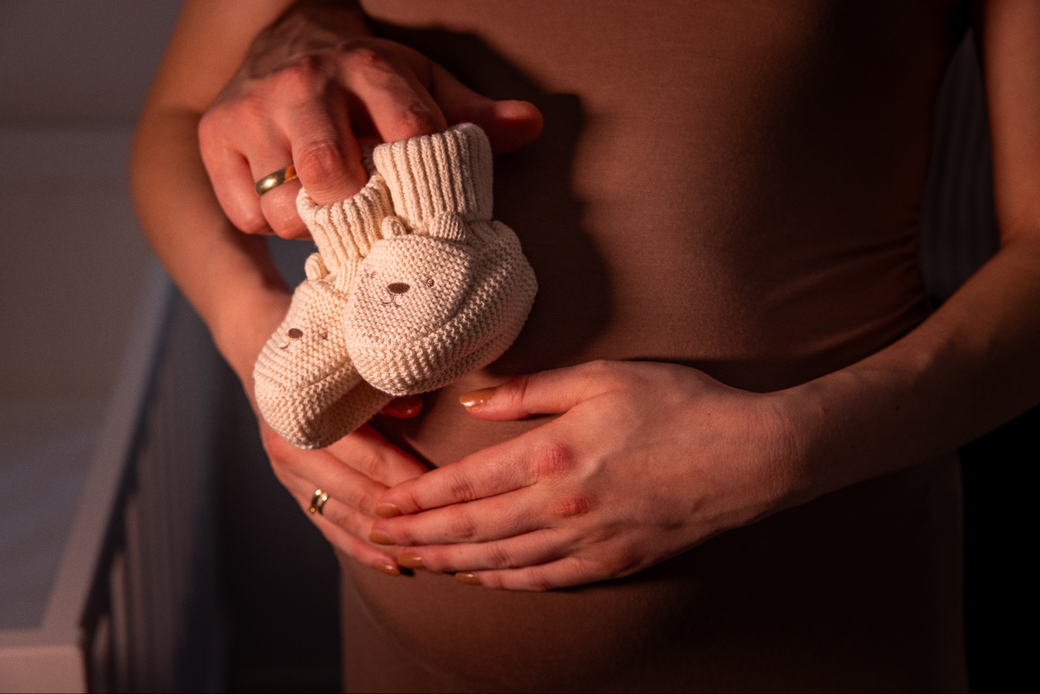 Zbliżenie na ciążowy brzuch kobiety, dłonie z obrączkami trzymają dziecięce, kremowe buciki z motywem misia. Delikatne oświetlenie podkreśla intymność chwili.
