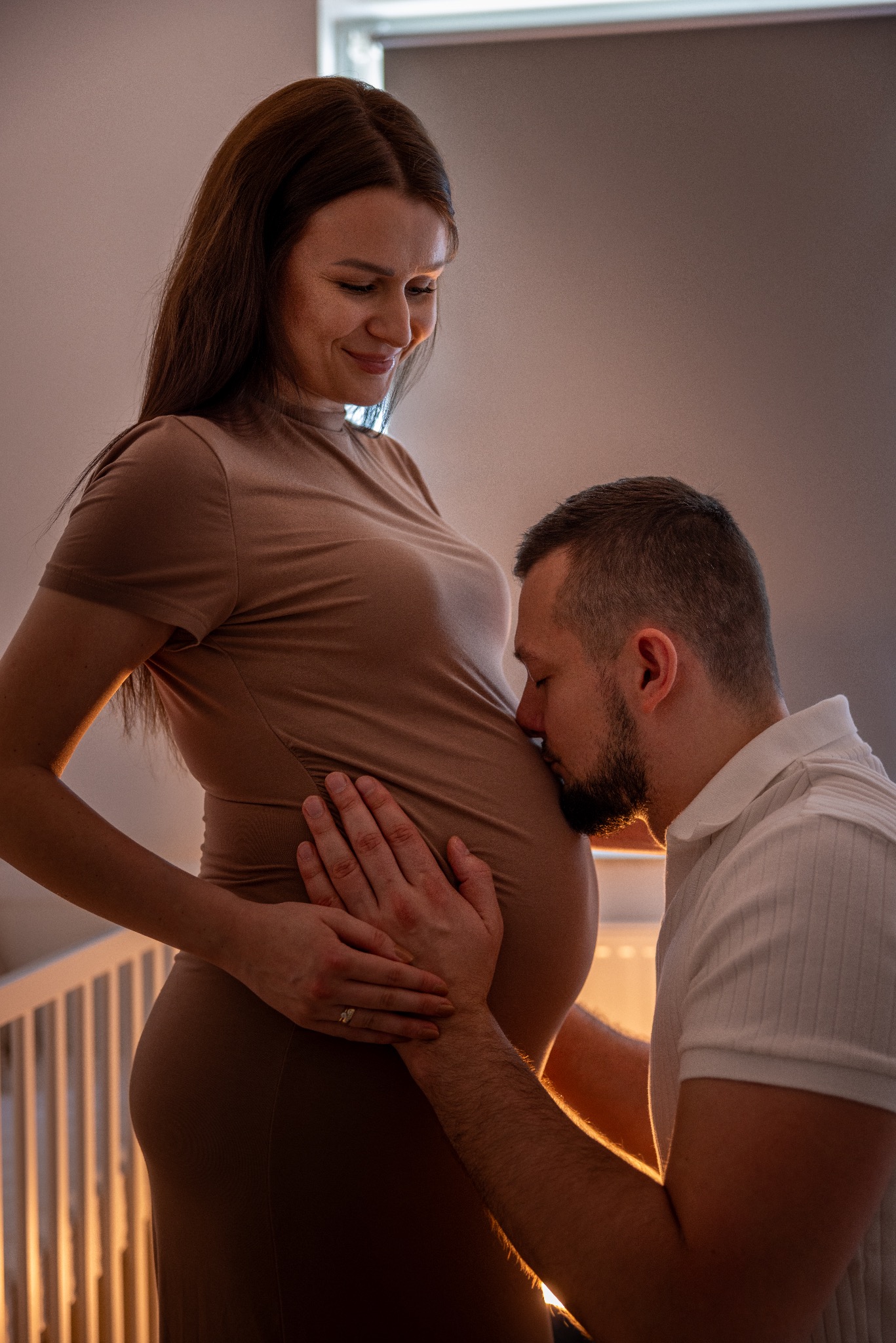 Czułe ujęcie: przyszła mama w brązowej sukience, tata całuje ciążowy brzuszek, dłoń na brzuchu. W tle białe łóżeczko dziecięce, ciepłe, intymne oświetlenie.