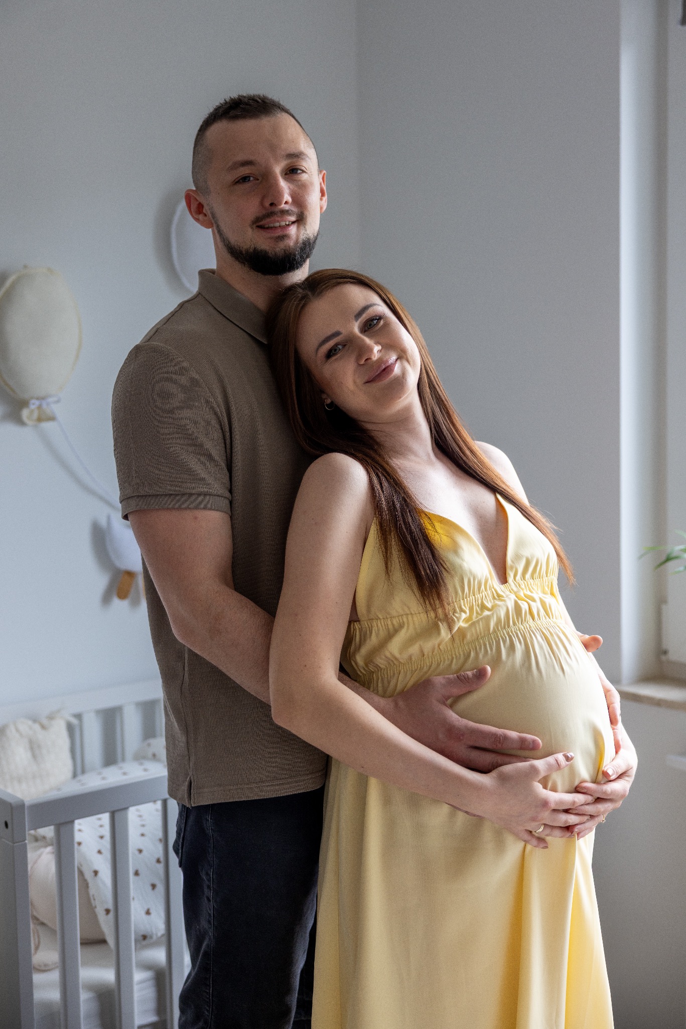 Uśmiechnięta para oczekująca dziecka, w jasnym pokoju z łóżeczkiem. Kobieta w żółtej sukience, mężczyzna obejmuje ją, dłonie na brzuchu. Delikatne światło.