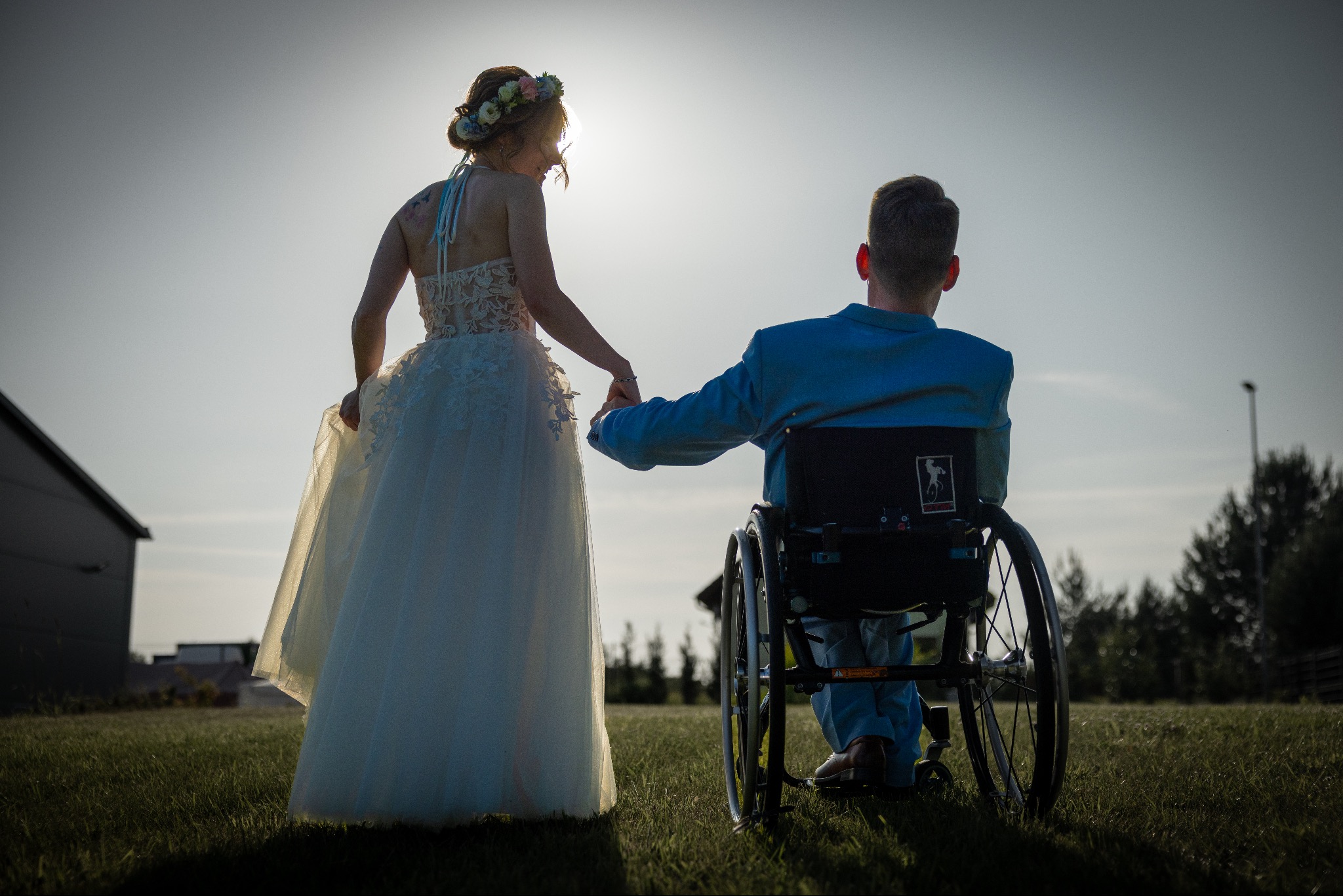 Para młoda na tle nieba: panna młoda w sukni ślubnej trzyma za rękę pana młodego na wózku inwalidzkim. Ujęcie z tyłu, oświetlenie kontrowe.
