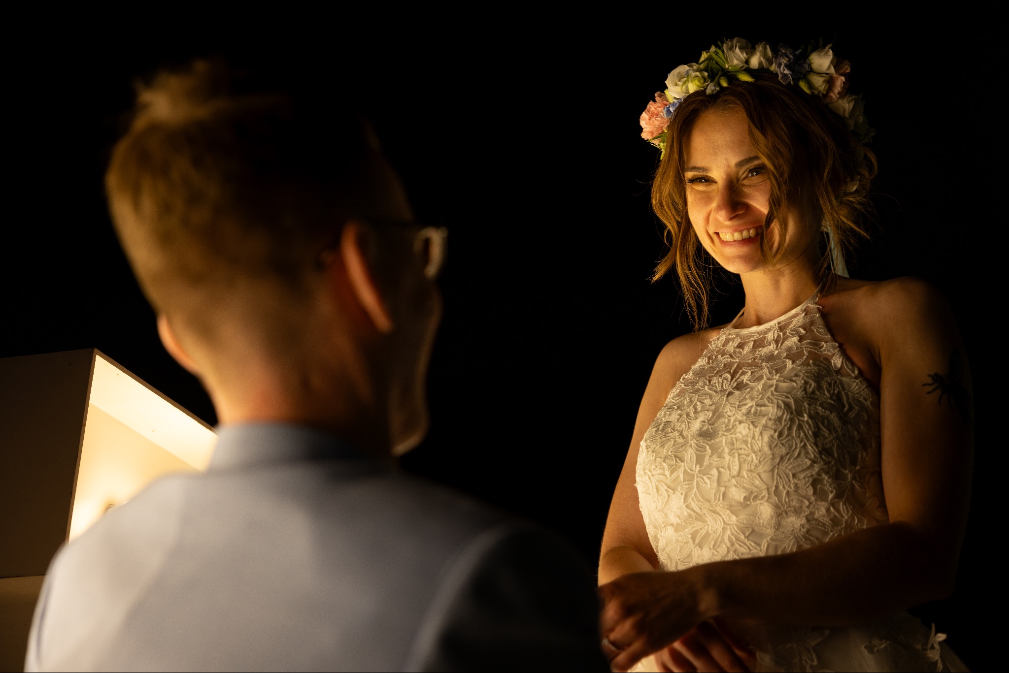 Uśmiechnięta panna młoda w wianku i koronkowej sukni, spogląda na mężczyznę w okularach, oświetlona ciepłym światłem na ciemnym tle.