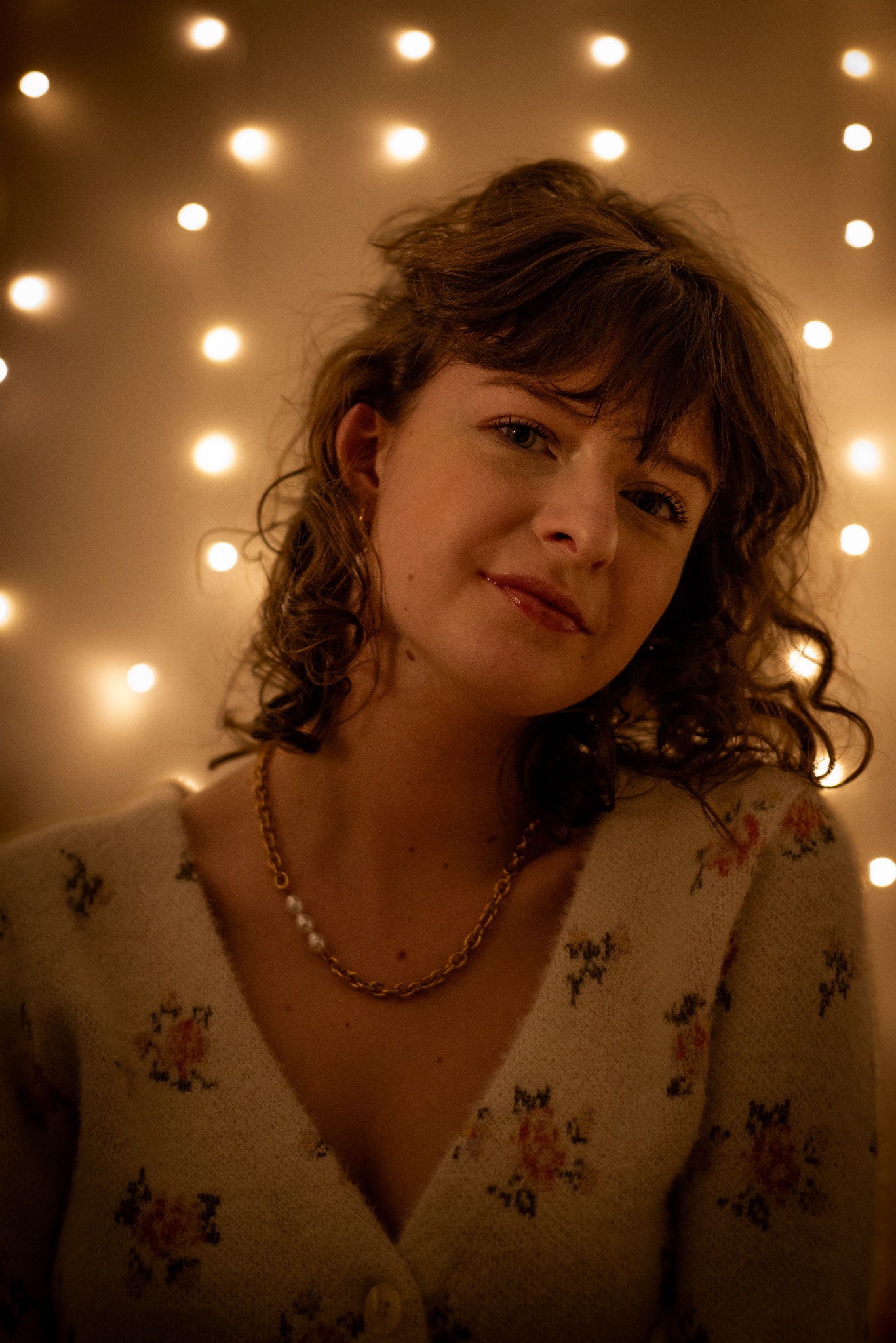 Portret młodej kobiety z delikatnym makijażem, w swetrze w kwiaty i złotej biżuterii, na tle ciepłych światełek, z lekko rozczochranymi włosami.