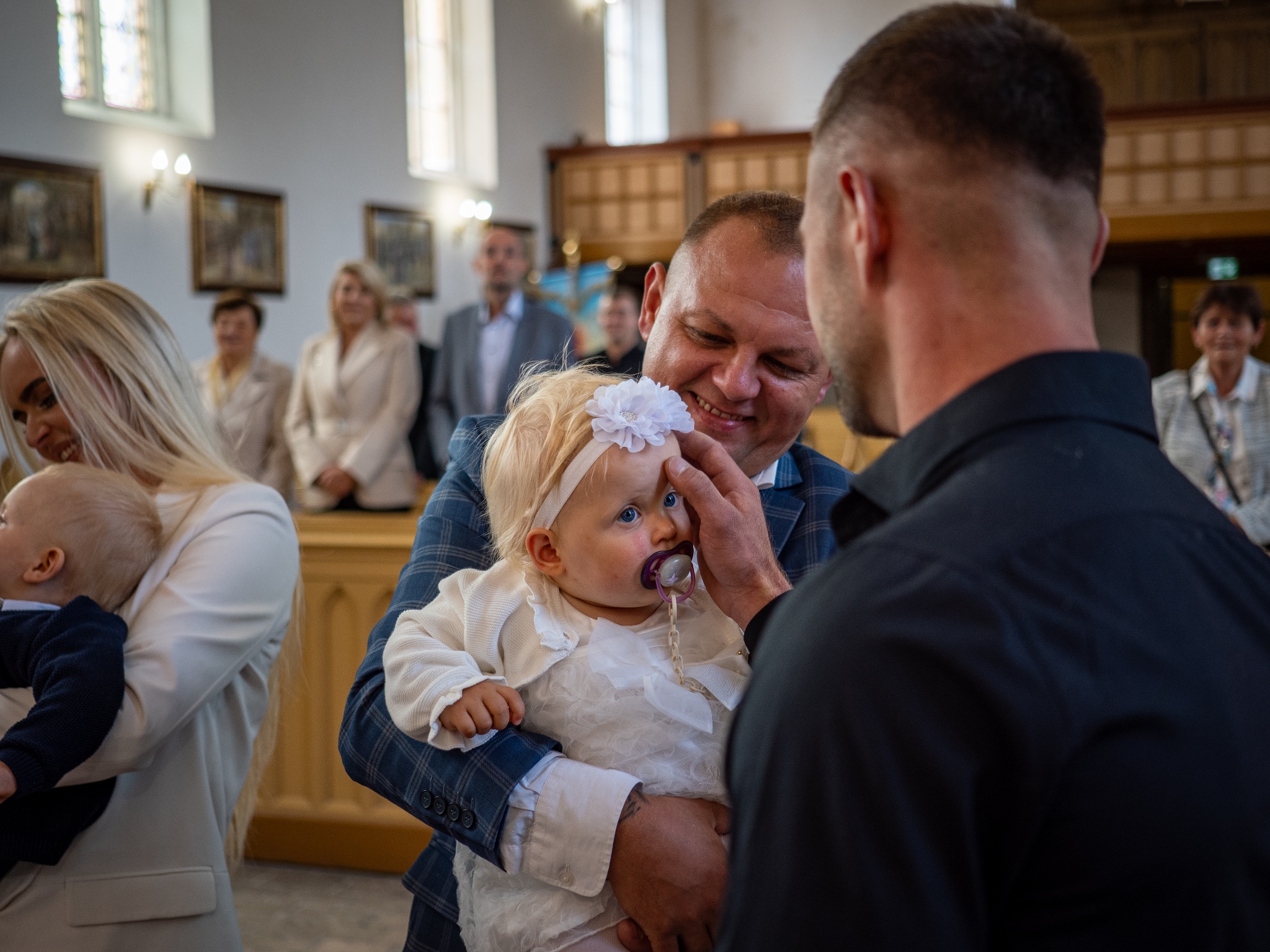 Uśmiechnięty mężczyzna w garniturze trzyma na rękach niemowlę w białej sukience i opasce z kwiatkiem, podczas uroczystości w kościele, w tle widoczni goście.
