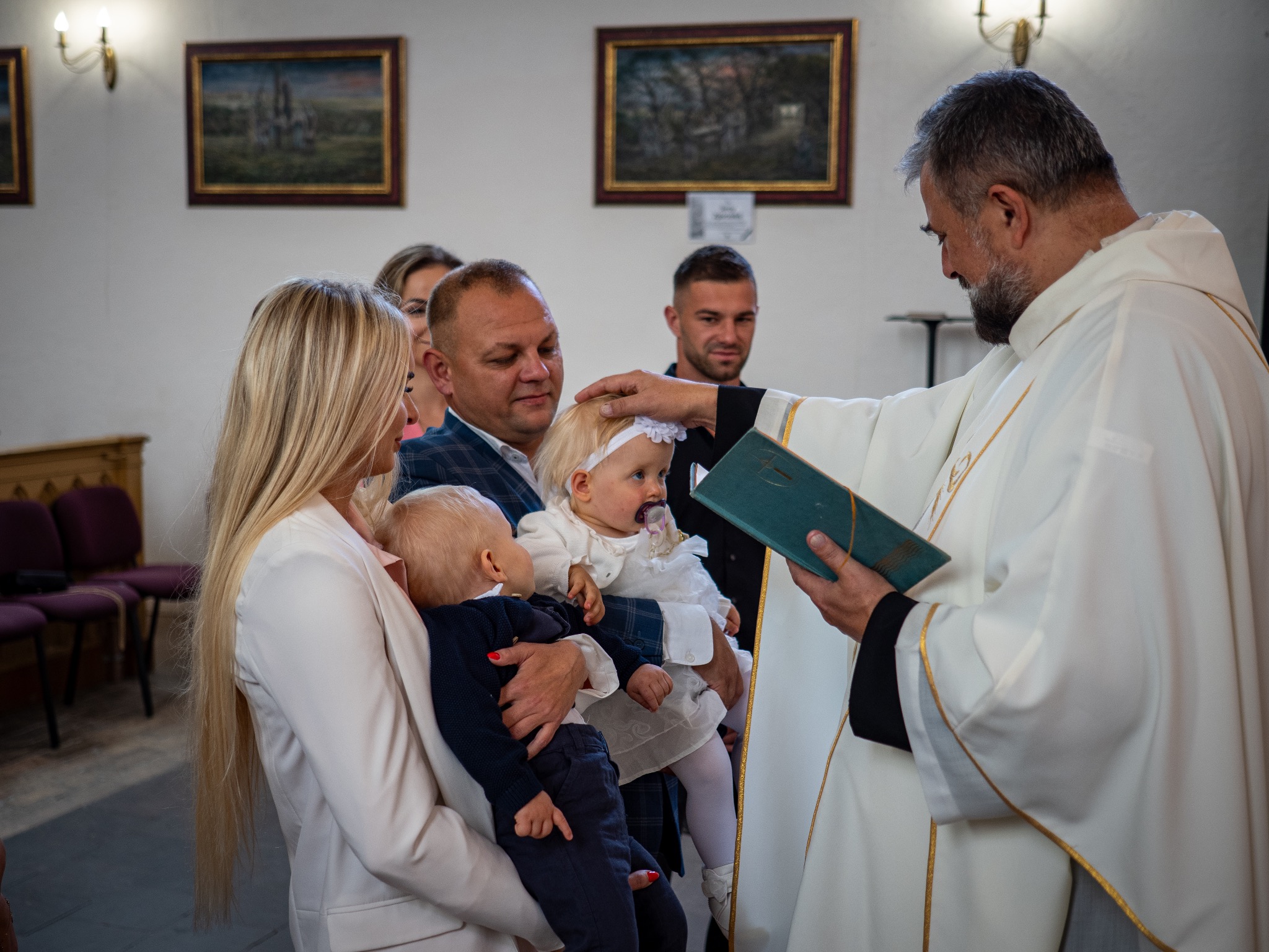Ksiądz udziela sakramentu chrztu dziewczynce w kościele. W tle rodzina dziecka. Uroczystość w jasnym wnętrzu z obrazami na ścianach.