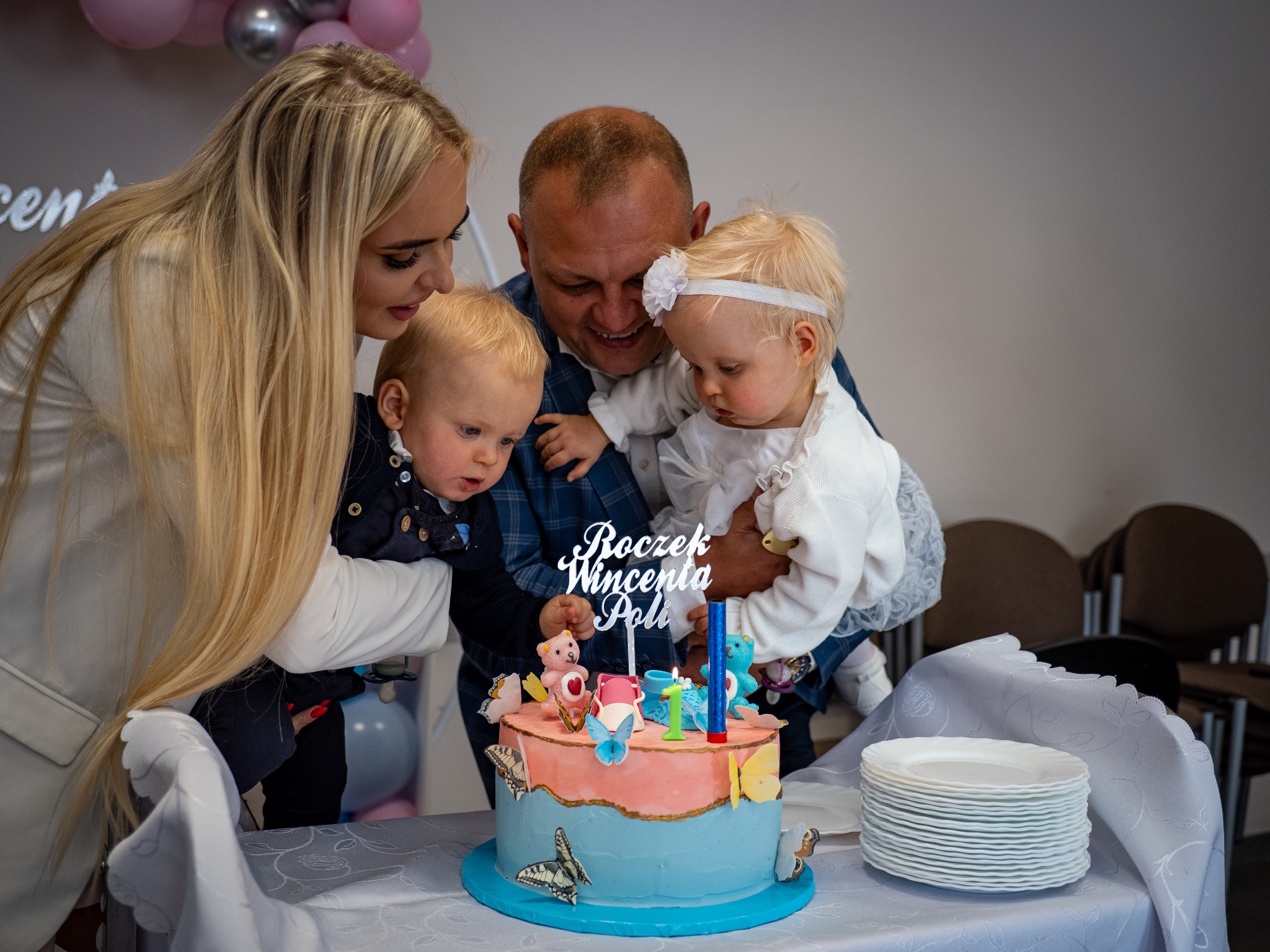 Rodzina świętuje pierwsze urodziny dziecka przy torcie z napisem 'Roczek Wincenta Poli', dekorowanego motylami i misiami. Dzieci obserwują płomień świeczki.