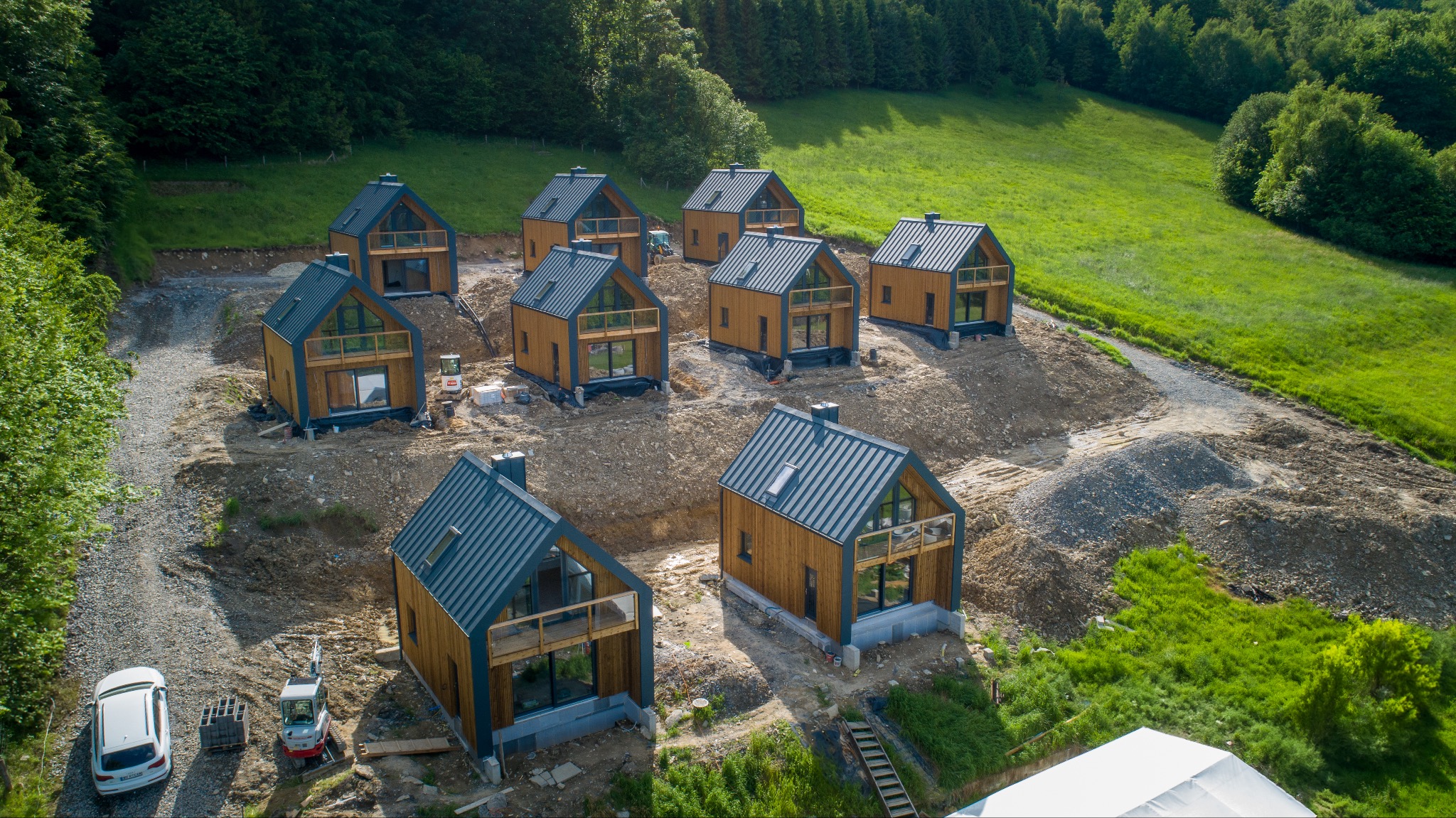 Widok z lotu ptaka na kompleks domków letniskowych z drewnianą elewacją i szarym dachem, usytuowanych na wzgórzu, w otoczeniu zieleni i z widocznymi maszynami budowlanymi.