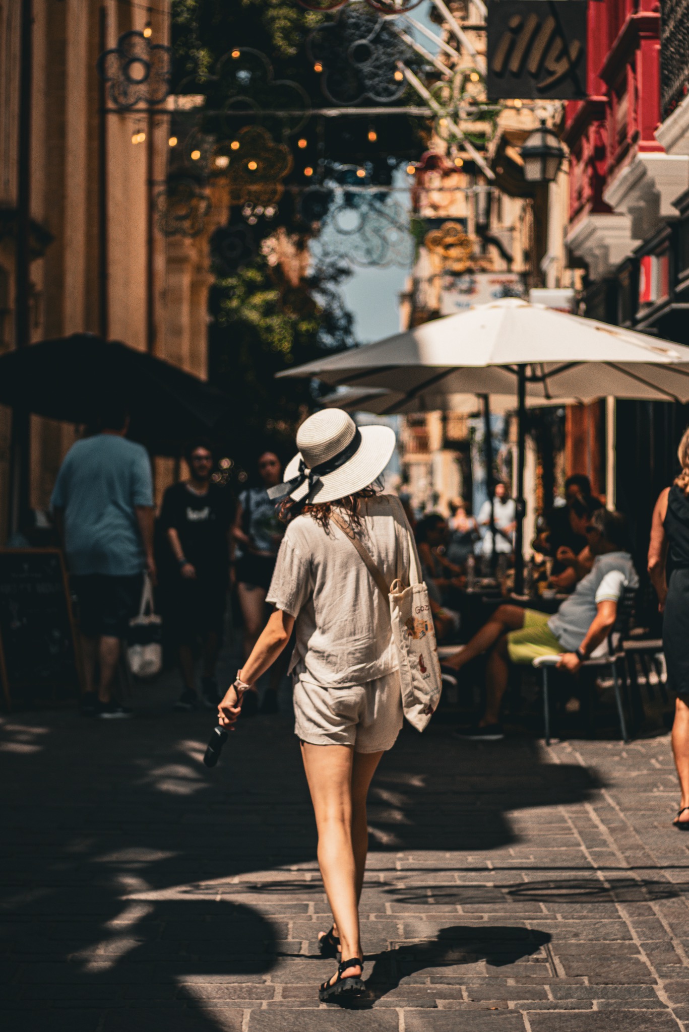 Kobieta w kapeluszu spaceruje po zacienionej uliczce z parasolami i stolikami kawiarnianymi w tle. Letni dzień, perspektywa z tyłu, naturalne światło.