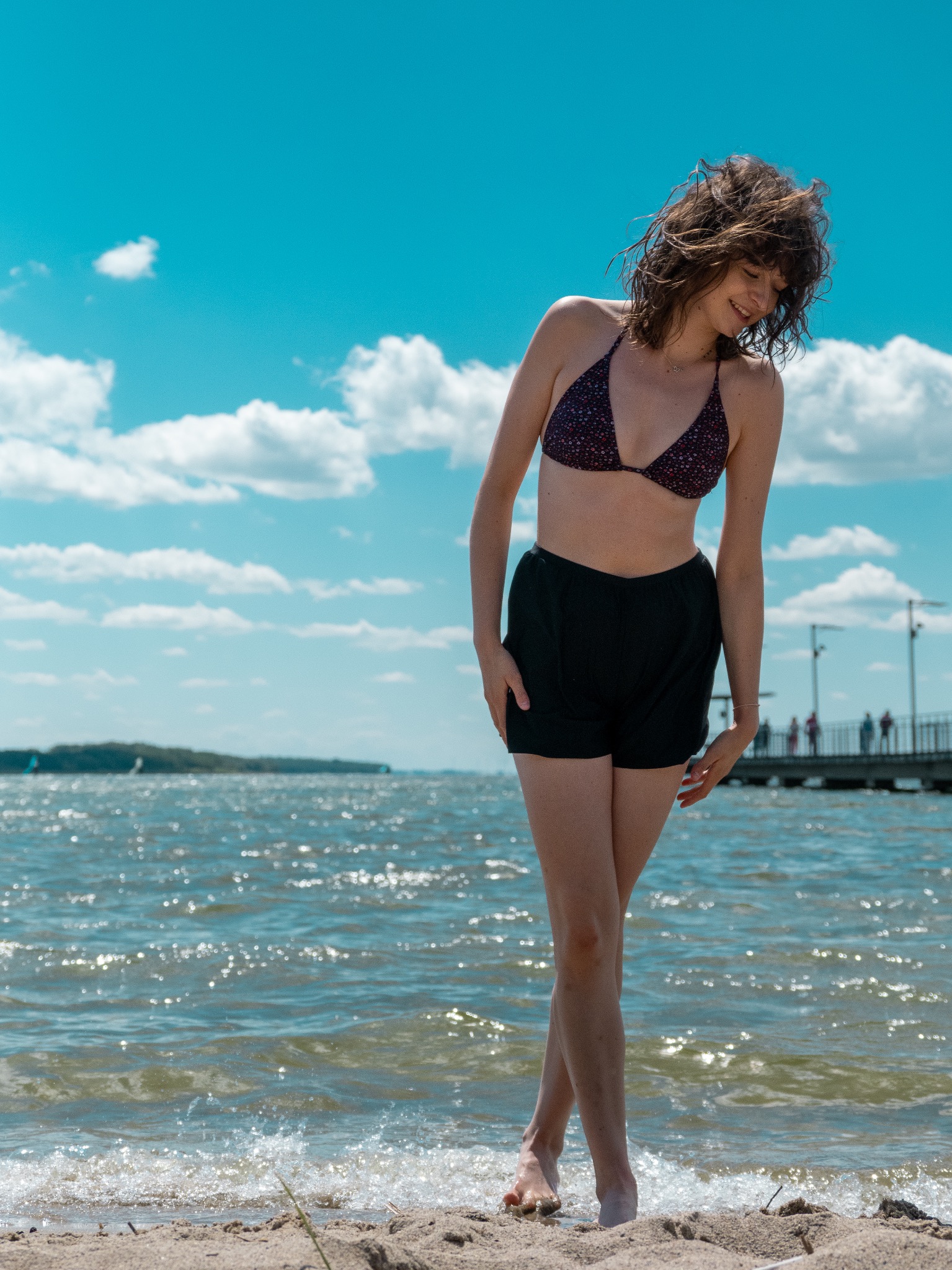 Młoda kobieta w bikini i czarnych szortach stoi na piaszczystej plaży, w tle woda i molo z ludźmi. Jasne niebo z chmurami, naturalne światło słoneczne.