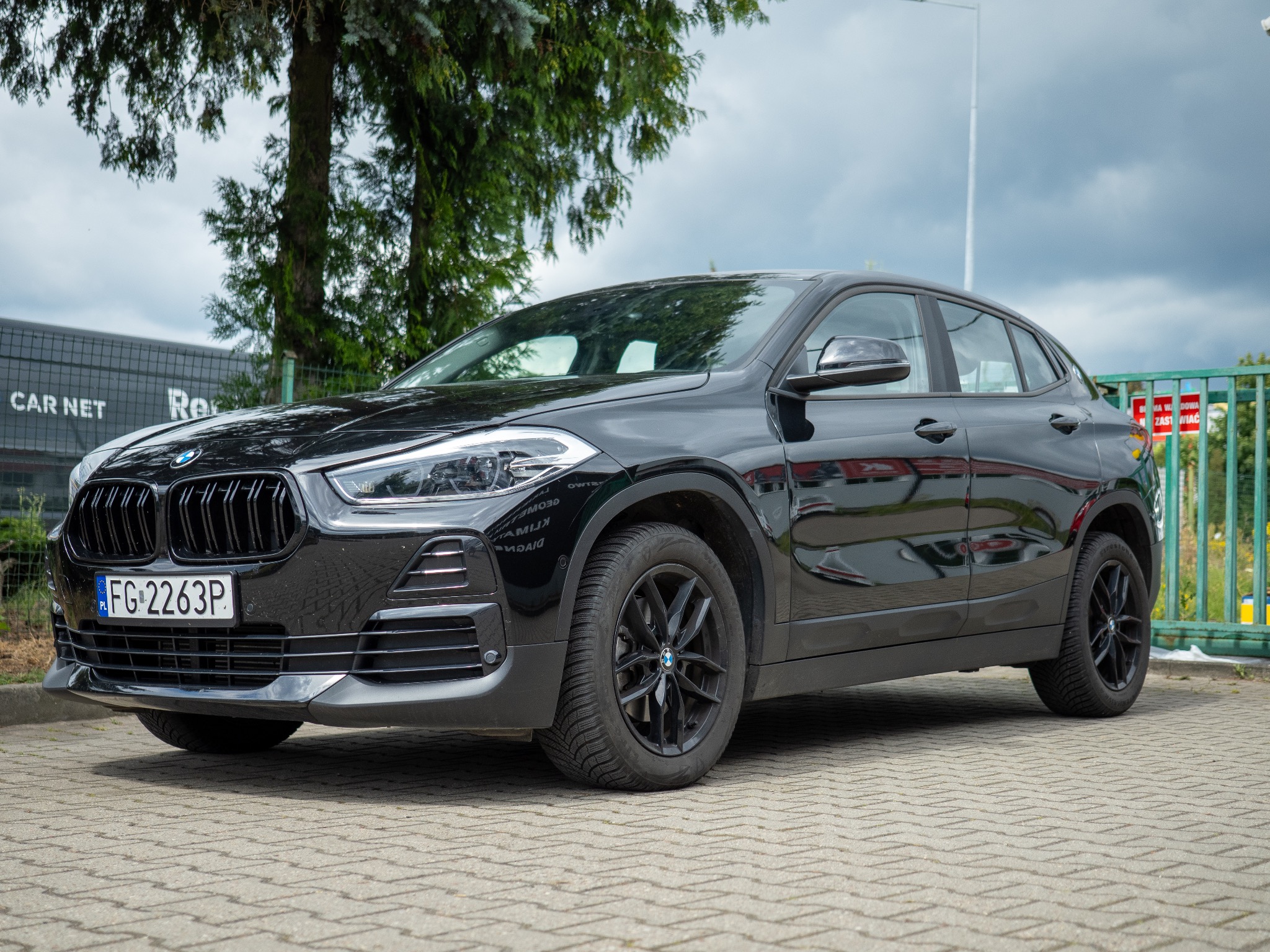 Czarny samochód BMW X2, model SUV, zaparkowany na brukowanej powierzchni. Widoczny z boku, z tablicą rejestracyjną FG 2263P. Tło stanowi zieleń drzew i budynek.