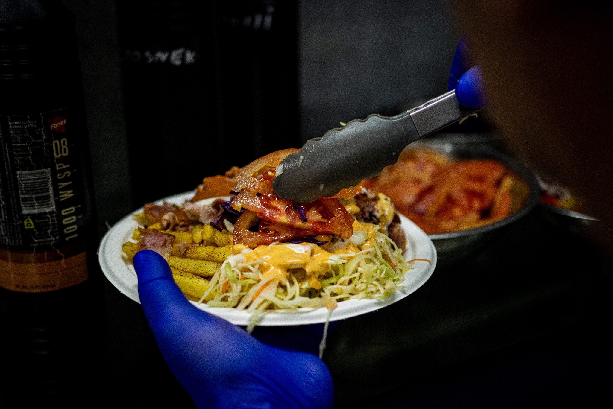 Nakładanie szczypcami pomidora na porcję frytek, surówki i mięsa na papierowym talerzu, trzymanym w dłoni w niebieskiej rękawiczce, w ciemnym otoczeniu.