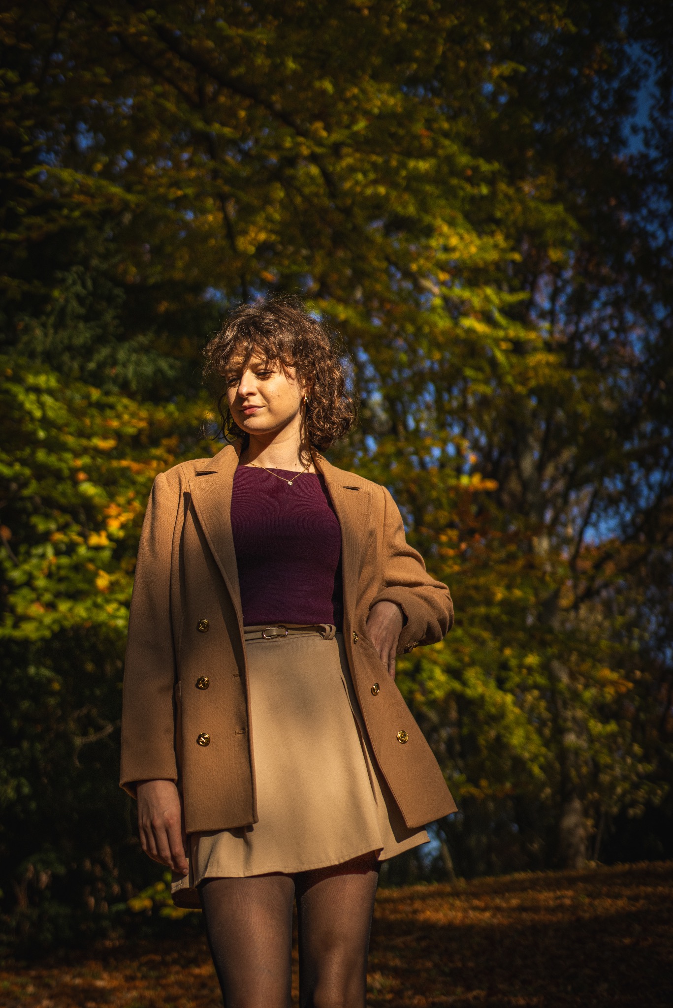 Portret kobiety w jesiennej scenerii, ubrana w płaszcz i spódnicę. Naturalne światło podkreśla ciepłe barwy liści w tle, tworząc klimatyczną atmosferę.