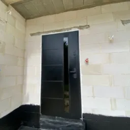 Czarne, nowoczesne drzwi zewnętrzne z szybą, zamontowane w surowym murze z białego bloczka. Widoczne zabezpieczenie hydroizolacyjne fundamentów.