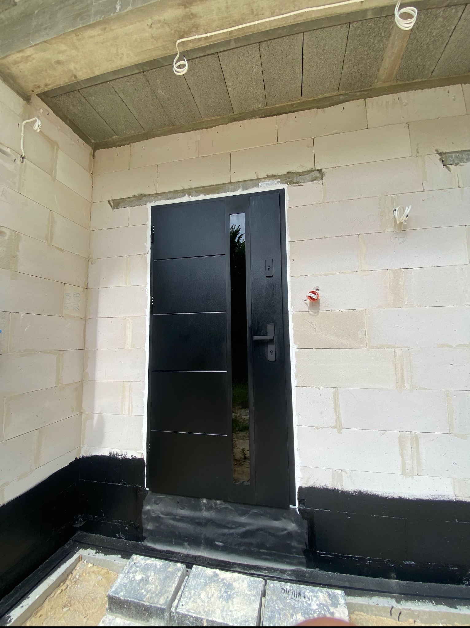 Czarne, nowoczesne drzwi zewnętrzne z szybą, zamontowane w surowym murze z białego bloczka. Widoczne zabezpieczenie hydroizolacyjne fundamentów.