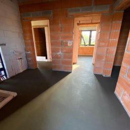 Michał Wójcik Domy ze Snów pl - Wnętrze w trakcie budowy: wylewka podłogowa, ściany z czerwonej cegły, widoczne instalacje sanitarne. Surowy stan, przygotowanie do dalszych prac wykończeniowych.
