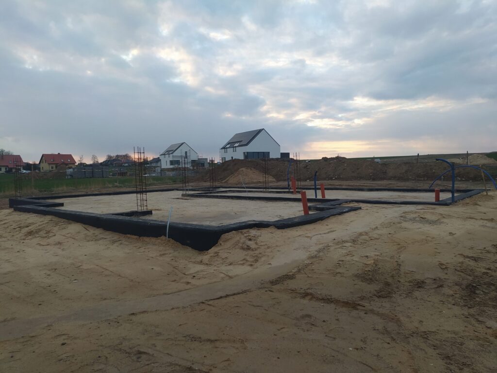 Fundament domu w trakcie budowy, zbrojenie, izolacja przeciwwilgociowa, rury instalacyjne. W tle domy jednorodzinne i zachmurzone niebo.