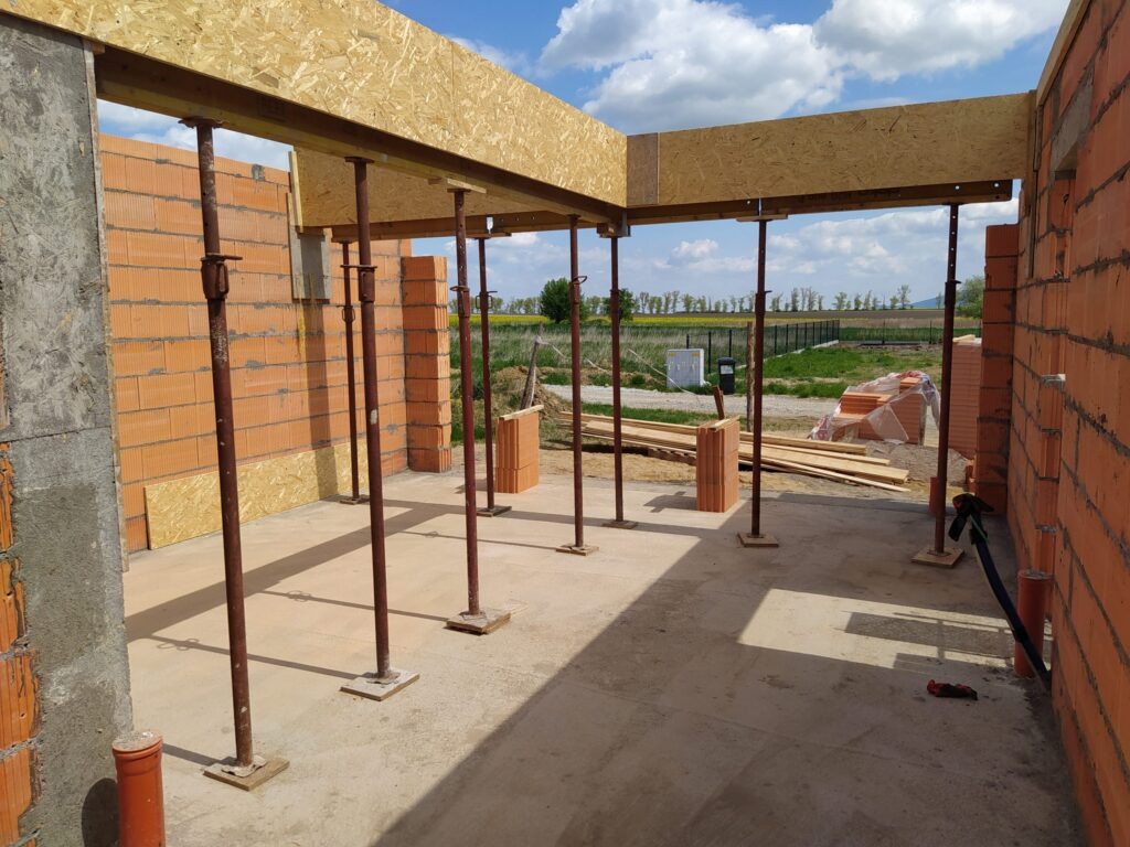 Wnętrze budowanego domu z czerwonej cegły. Widoczne podpory stropowe, drewniane elementy szalunku i częściowo wymurowane ściany. W tle krajobraz.