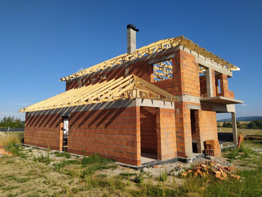 Niedokończony dom z czerwonej cegły z drewnianą konstrukcją dachu na tle błękitnego nieba. Widoczne niezabezpieczone otwory okienne i drzwiowe.