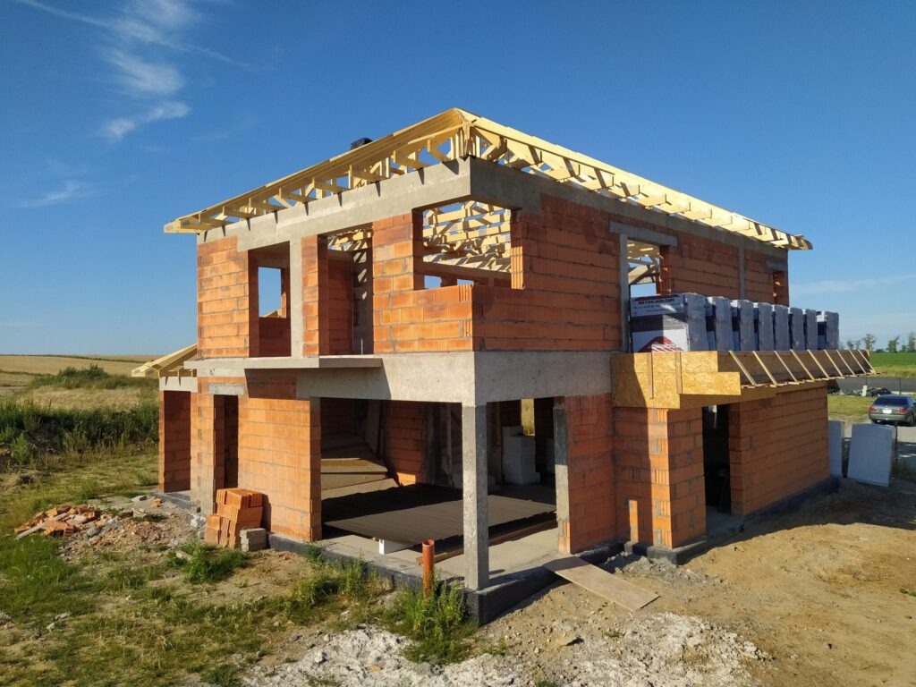 Dwukondygnacyjny dom w trakcie budowy, z czerwonej cegły, z widocznym drewnianym szkieletem dachu i materiałami budowlanymi na balkonie, na tle błękitnego nieba.