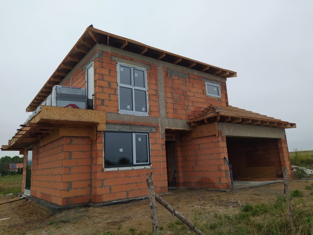 Surowy dom z czerwonej cegły w trakcie budowy, z zamontowanymi oknami i częściowo wykonanym dachem, widok z zewnątrz w pochmurny dzień.