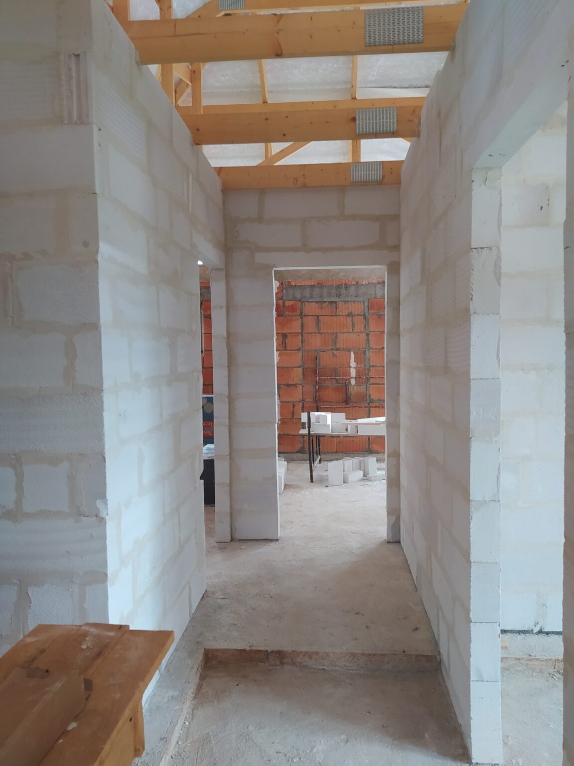 Wnętrze w trakcie budowy: białe ściany z betonu komórkowego, widoczne drewniane elementy konstrukcji dachu i fragment ściany z czerwonej cegły w głębi. Surowy stan, brak wykończenia.