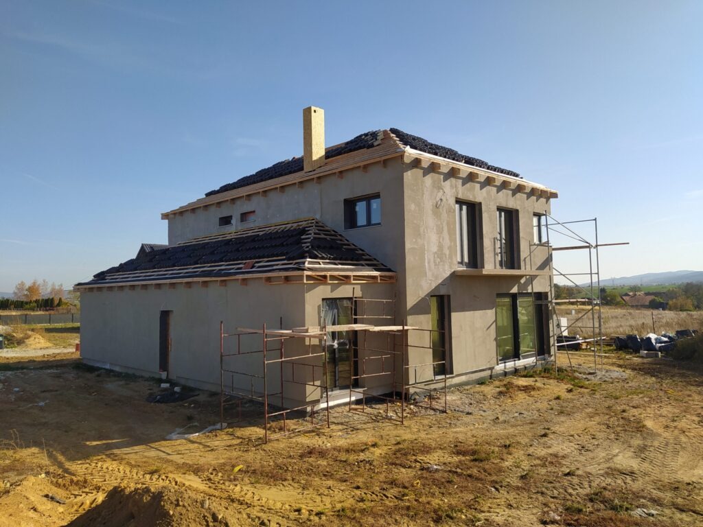 Surowy dom z ciemną dachówką w trakcie budowy, z widocznymi rusztowaniami i elementami konstrukcyjnymi dachu. Tło stanowi błękitne niebo i odległy krajobraz.