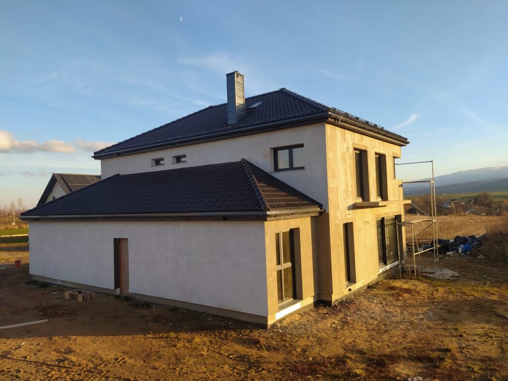 Dwukondygnacyjny dom w stanie surowym zamkniętym z ciemną dachówką, widok z boku na tle krajobrazu z górami w oddali i rusztowaniem przy ścianie.