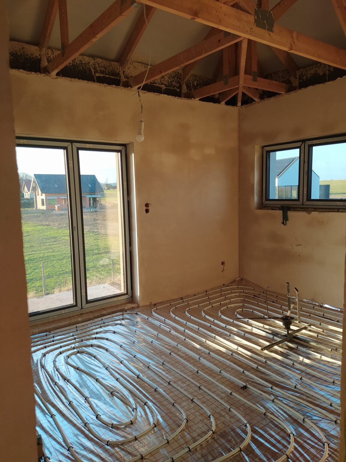 Wnętrze w trakcie budowy: surowe ściany, okna z widokiem na domy, instalacja ogrzewania podłogowego na folii, drewniana konstrukcja dachu z wiszącą żarówką.