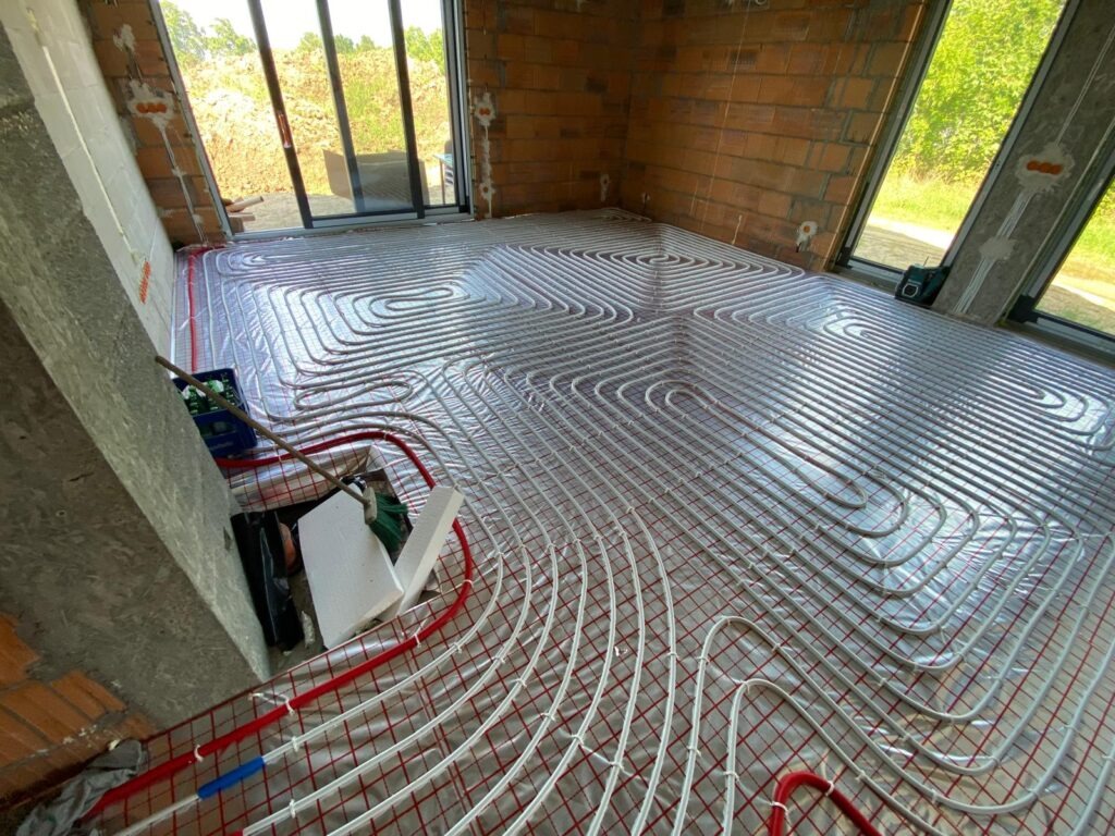 Instalacja ogrzewania podłogowego w surowym wnętrzu z ceglanymi ścianami i dużymi oknami. Widoczne białe rury ułożone w pętle na folii izolacyjnej.