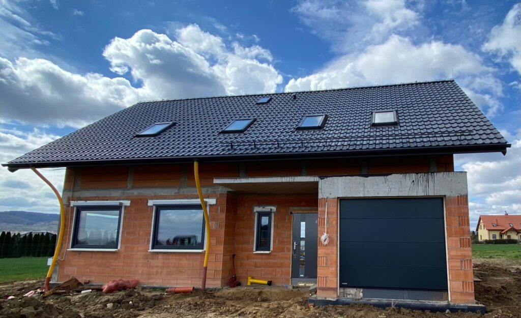 Niedokończony dom z czerwonej cegły z czarnym dachem i oknami dachowymi, widok z zewnątrz, w tle zielone pola i błękitne niebo z chmurami.