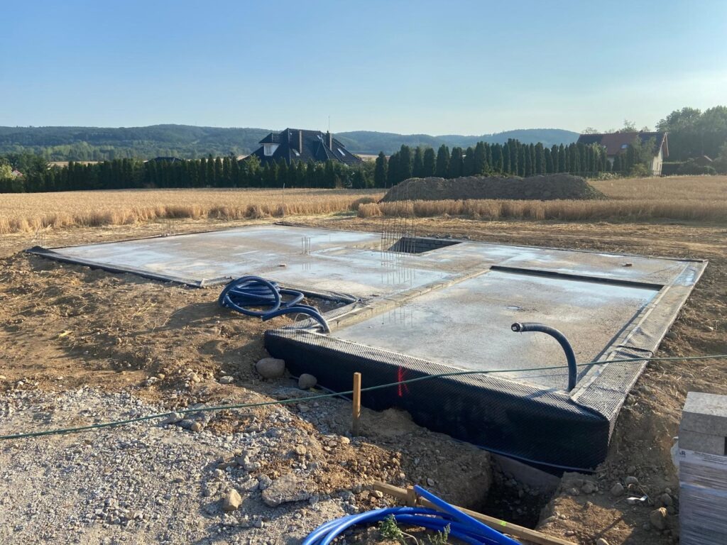 Wylana płyta fundamentowa z hydroizolacją na działce budowlanej, widok na pola i dom w tle. Etap budowy domu jednorodzinnego.
