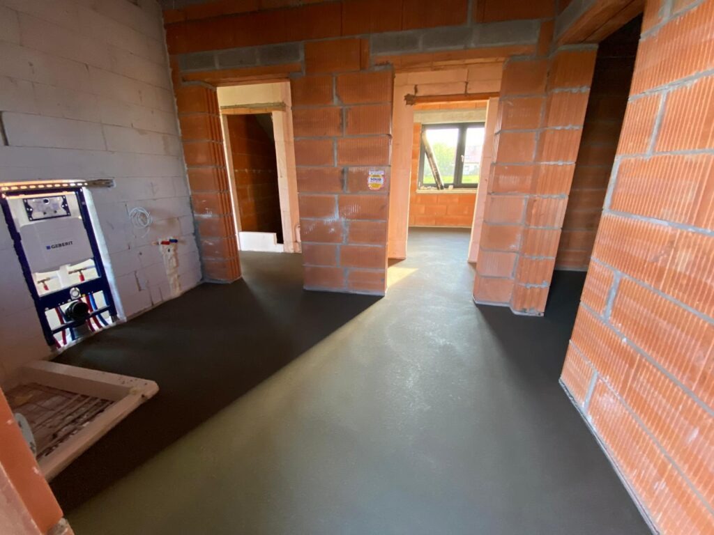 Wnętrze w trakcie budowy: wylewka podłogowa, ściany z czerwonej cegły, widoczne instalacje sanitarne. Surowy stan, przygotowanie do dalszych prac wykończeniowych.